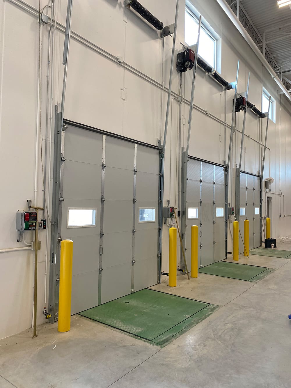 Three industrial garage doors with small windows are lined up inside a warehouse. Yellow safety bollards and green metal plates protect each door, reflecting the organized efficiency valued by American business. The area is well-lit with high ceilings and white walls.