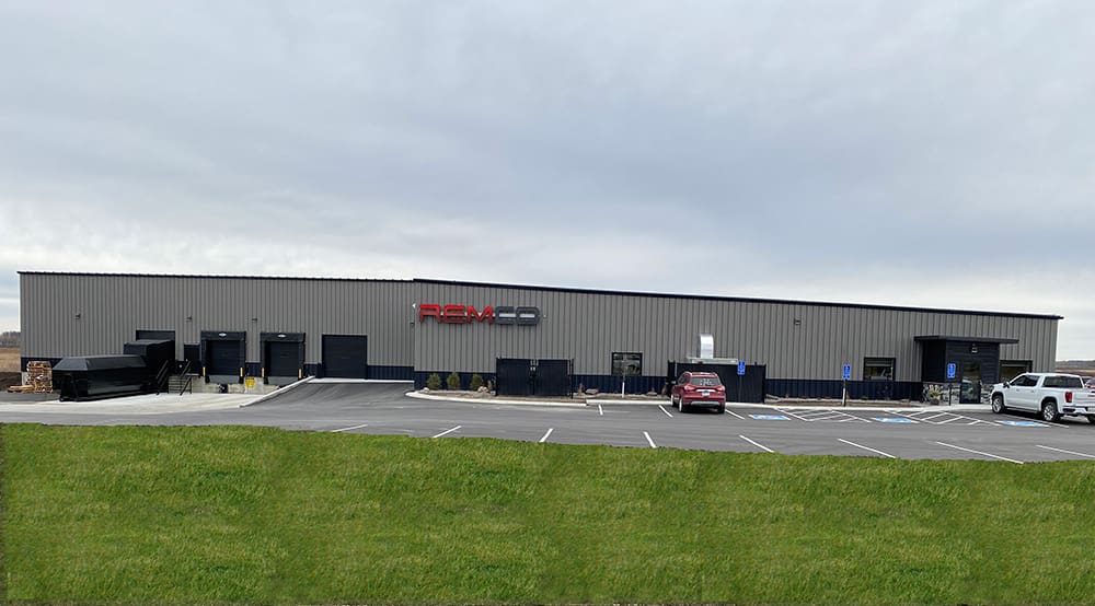 A wide, modern industrial building with a gray exterior and the red and black "REMCO" sign above the entrance, surrounded by a parking lot with several cars and grassy foreground.