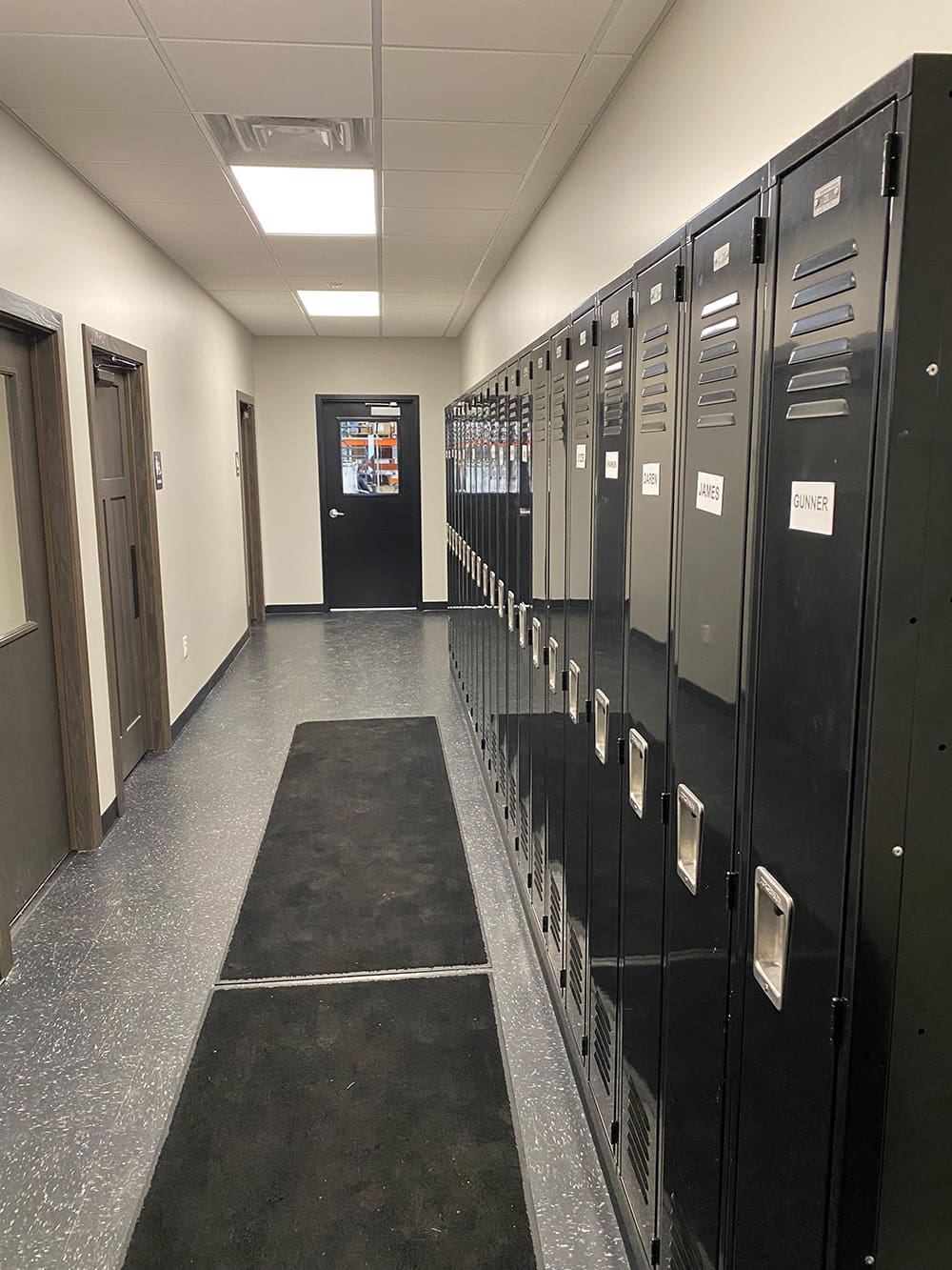A hallway with black lockers on the right, each labeled with a name. On the left are several doors, and at the end of the hallway is a closed door with a window. The floor is gray with black mats.
