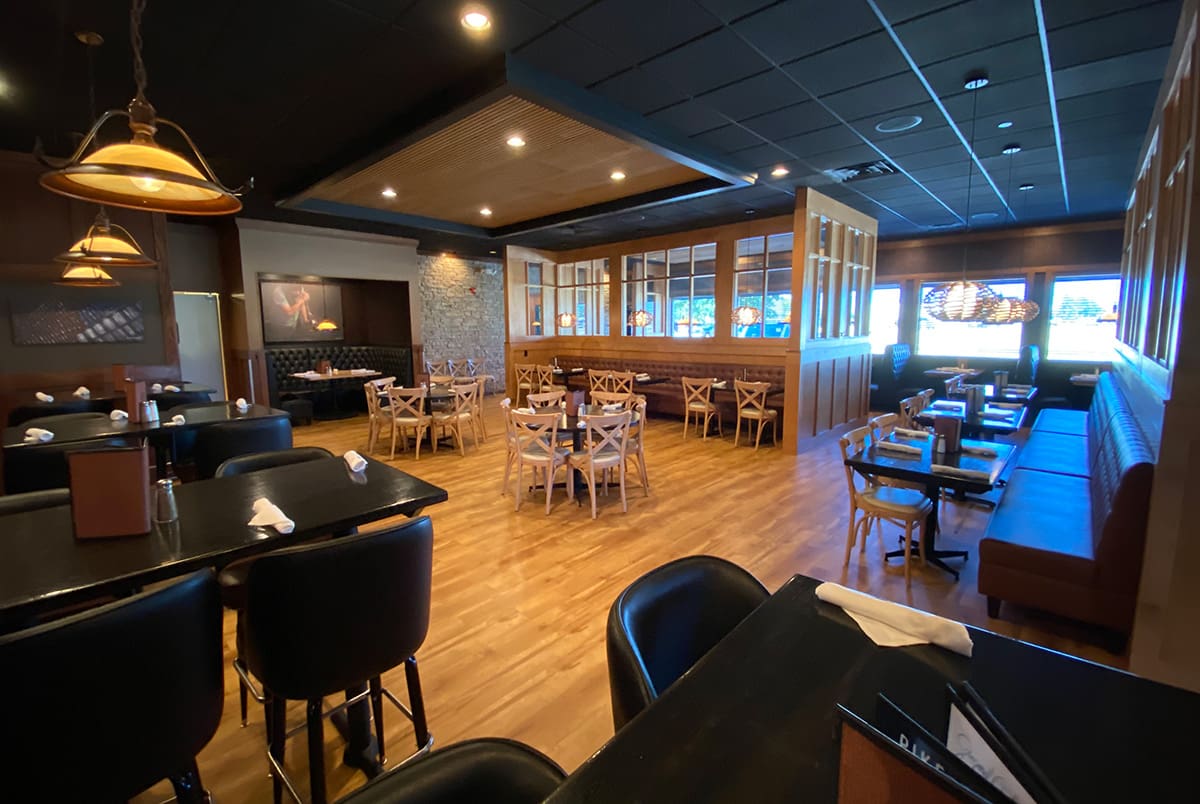 A spacious, modern restaurant with wooden floors, tables, and chairs. There are black leather chairs, booth seating along the windows, and warm overhead lighting. The dining area is empty and neatly arranged.
