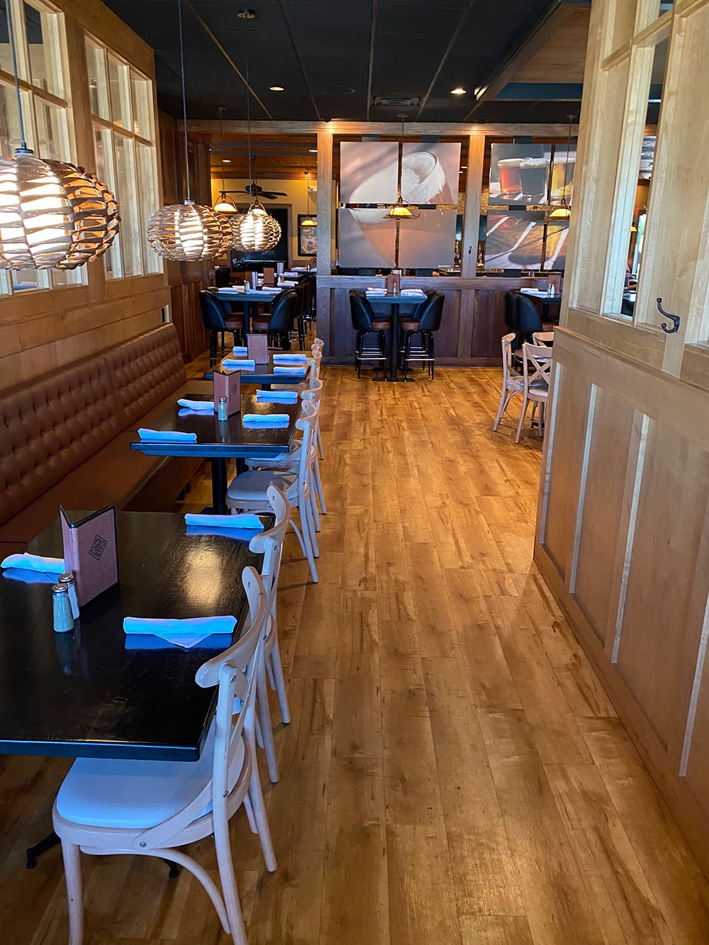 A cozy restaurant interior with wooden floors, tables set with blue napkins, cushioned bench seating on one side, and pendant lights hanging above. More tables and seating are visible in the background.