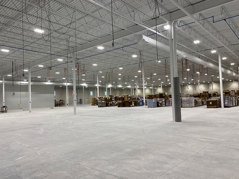 A spacious warehouse in the H. Boyd Addition with high ceilings, bright overhead lighting, and concrete floors. Pallets with boxes and goods are stacked along the back wall, while the foreground is mostly empty.