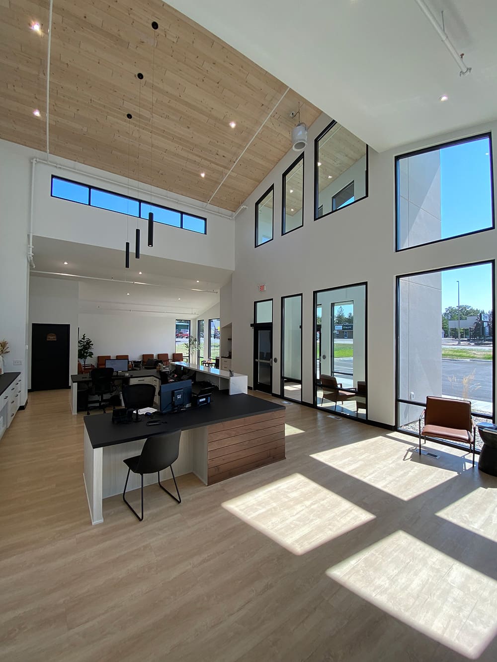 Modern office lobby with high ceilings, large windows letting in natural light, a black reception desk, seating area with brown chairs, light wood floors, and white walls welcomes you to Woodland Dental.