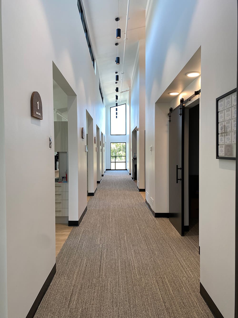 A modern hallway with high ceilings, carpeted floor, several open doorways on both sides, and a large window at the end letting in natural light welcomes patients into Woodland Dental’s inviting dental clinic.