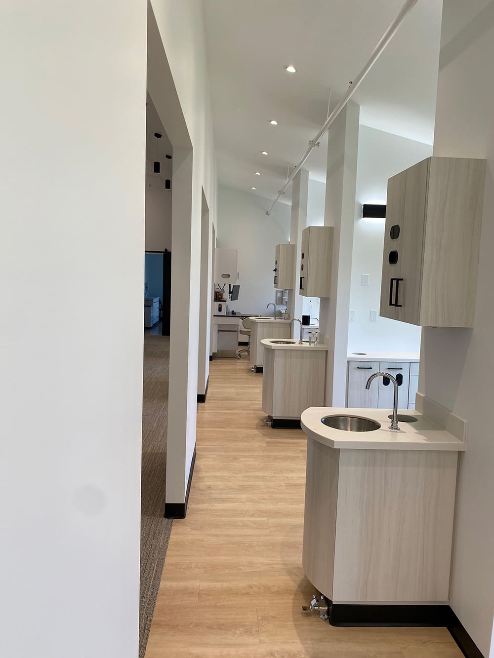 A modern Woodland Dental clinic interior with light wood flooring, white walls, and multiple dentist treatment stations separated by partial walls. Each area features sinks and cabinets, creating a clean and brightly lit space.
