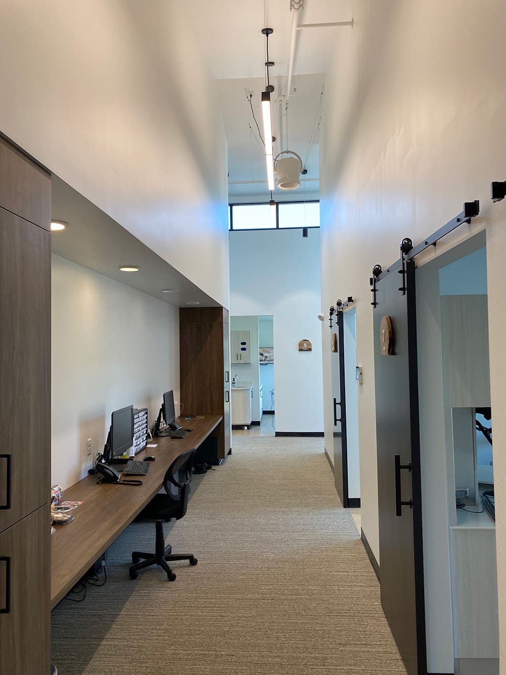 A modern office hallway at Woodland Dental features a long wooden desk with two office chairs and computers on the left, sliding barn doors on the right, and bright natural light streaming in from a high window at the end.