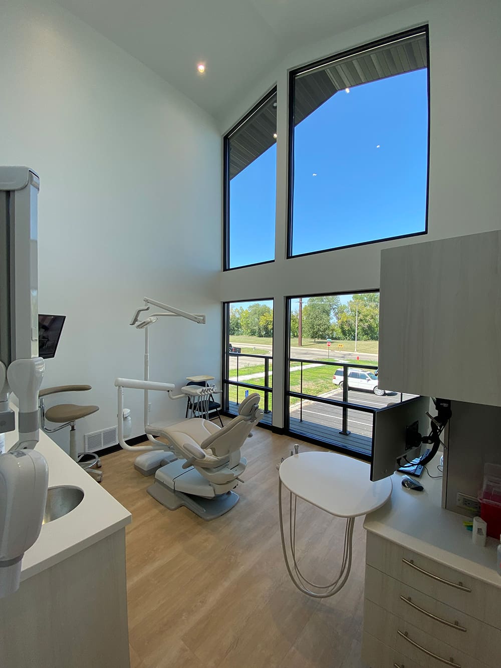 A modern Woodland Dental clinic featuring a reclining exam chair, advanced dental equipment, light wood floors, tall ceilings, and large windows showcasing green trees and a blue sky outside.