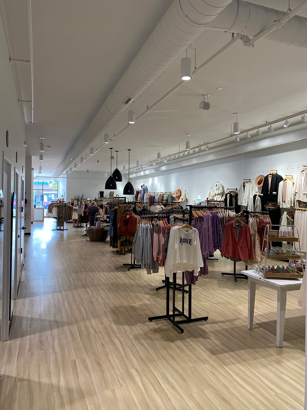 Creative Touch Boutique is a brightly lit clothing store with light wood floors, racks of colorful clothes, hats, and jewelry displays. Pendant lights hang from the ceiling as daylight shines through large front windows.