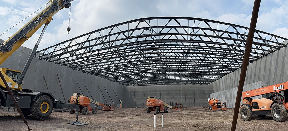 Construction site of a large building with steel roof beams, high concrete walls, cranes, and orange construction vehicles under a partly cloudy sky.